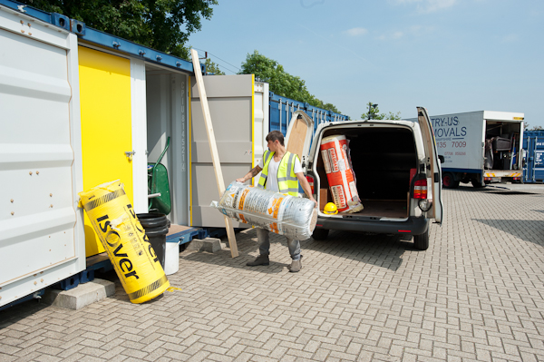 Man in a high-vis vest carrying insulation rolls from a van next to a storage container.